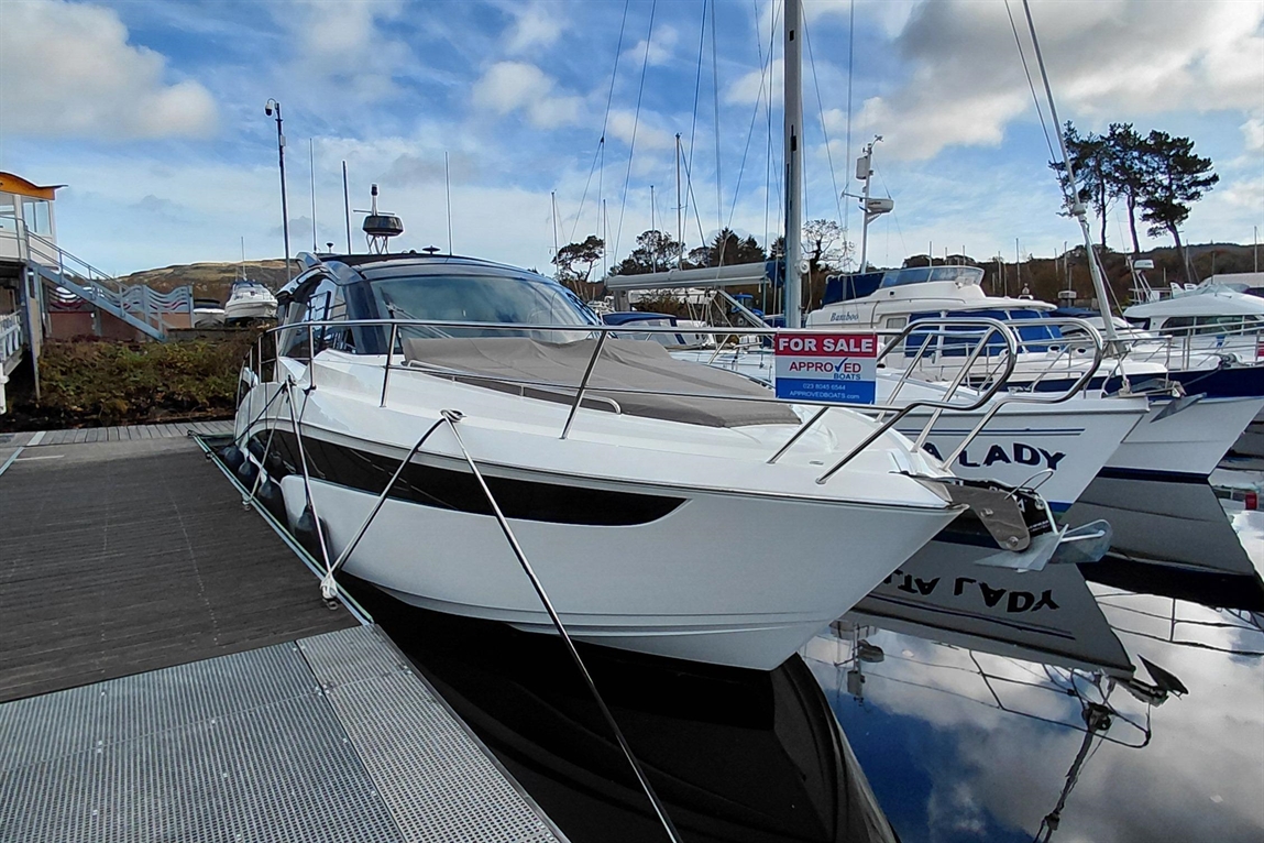 A sleek white boat docked at a marina with a "FOR SALE" sign, surrounded by other vessels under a cloudy sky.